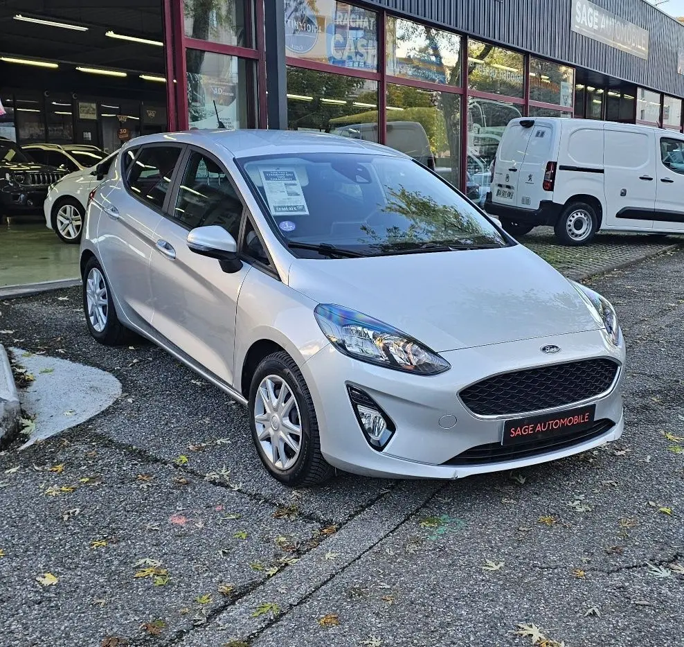 Ford Fiesta 2021 gris lunaire métal vue 3/4 avant droit, avec calandre noire et phares allumés devant un garage Sage Automobile.