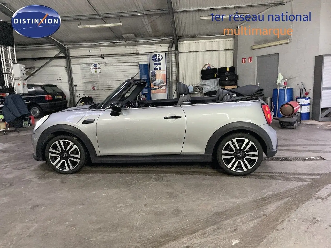 Profil droit d'une MINI Cabriolet 2023 couleur Melting Silver Metal avec capote ouverte dans un garage.