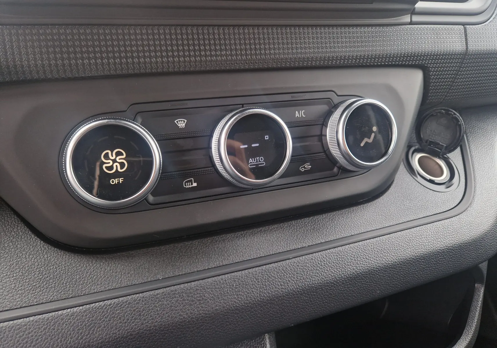 Gros plan sur les commandes de climatisation du Renault Trafic Fourgon gris urbain 2025, avec boutons ronds et prise allume-cigare ouverte.