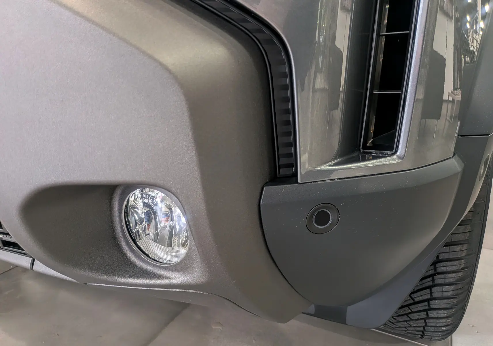 Gros plan sur le pare-chocs avant droit gris schiste du Dacia Duster 2025, avec antibrouillard et radar de stationnement visible.