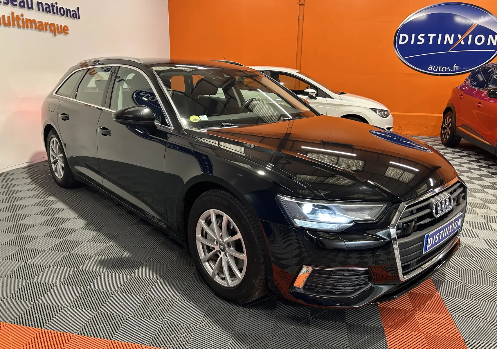 Audi A6 Avant noire vue en 3/4 avant droit, phares LED allumés, dans un showroom avec sol damier.