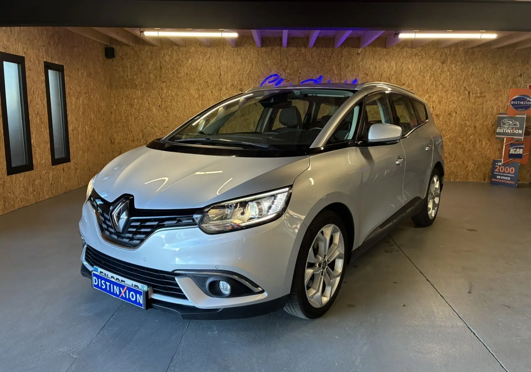 Renault Grand Scenic gris vue 3/4 avant droit dans un garage avec éclairage intérieur et plaques Distinxion.