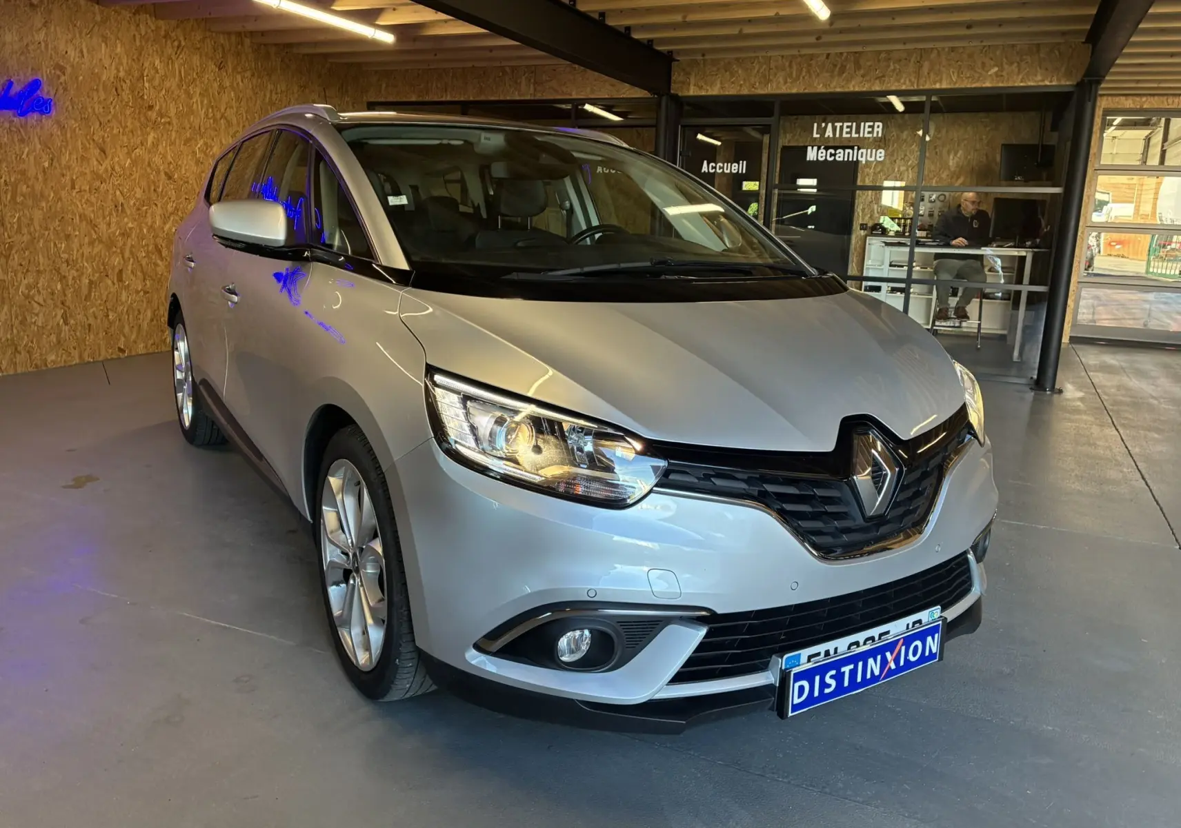 Renault Grand Scenic gris en 3/4 avant droit, stationné dans un atelier mécanique avec éclairage intérieur.