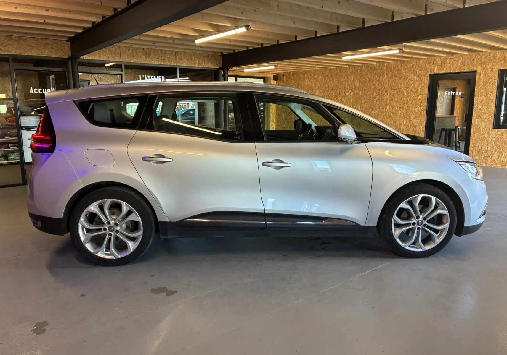 Profil côté gauche d'un Renault Grand Scenic gris 2017 en intérieur, avec ses jantes alliage et ses vitres teintées.