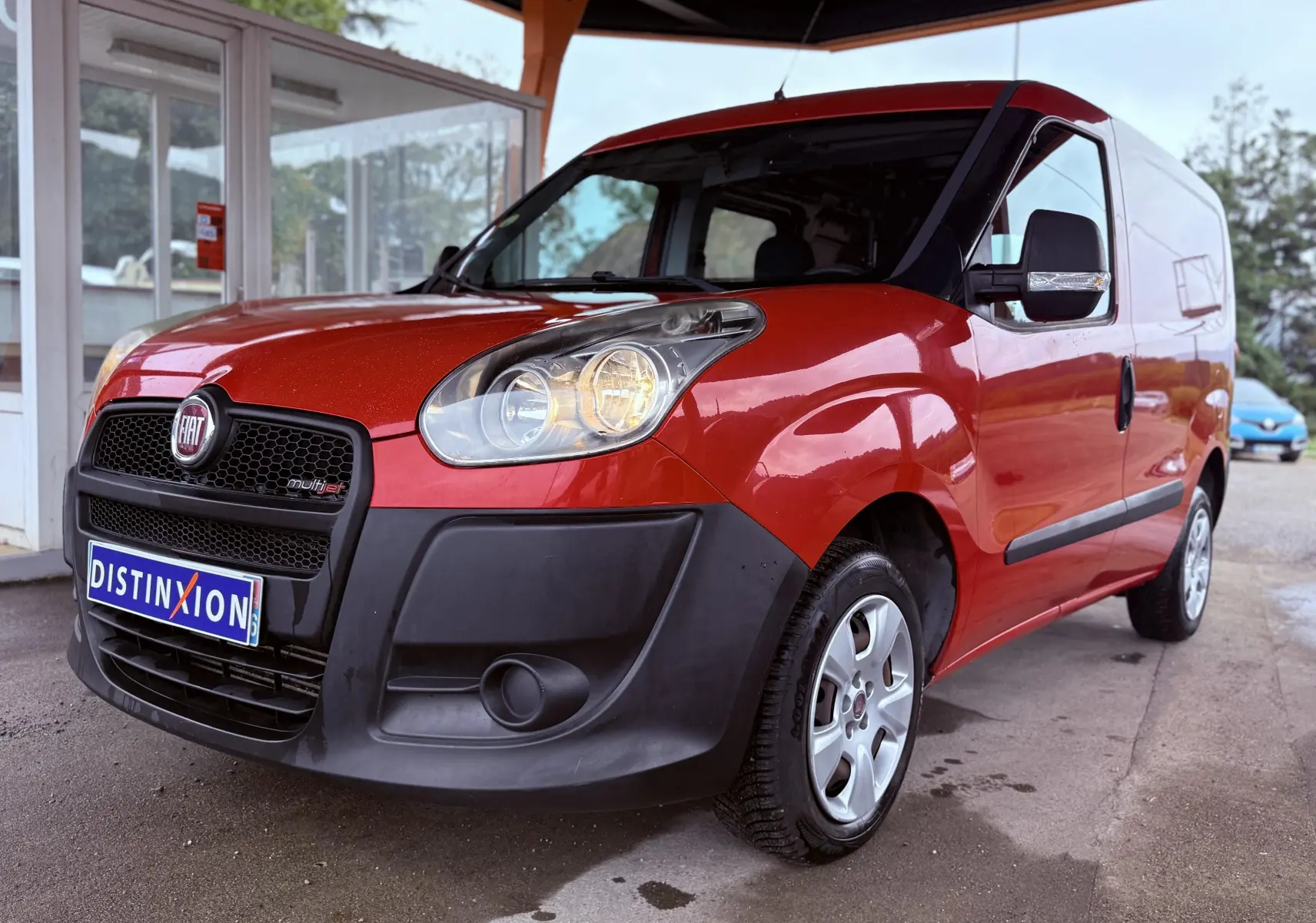 Fiat Doblo Cargo rouge vu en 3/4 avant droit, avec pare-chocs noir et phares allumés sous abri.