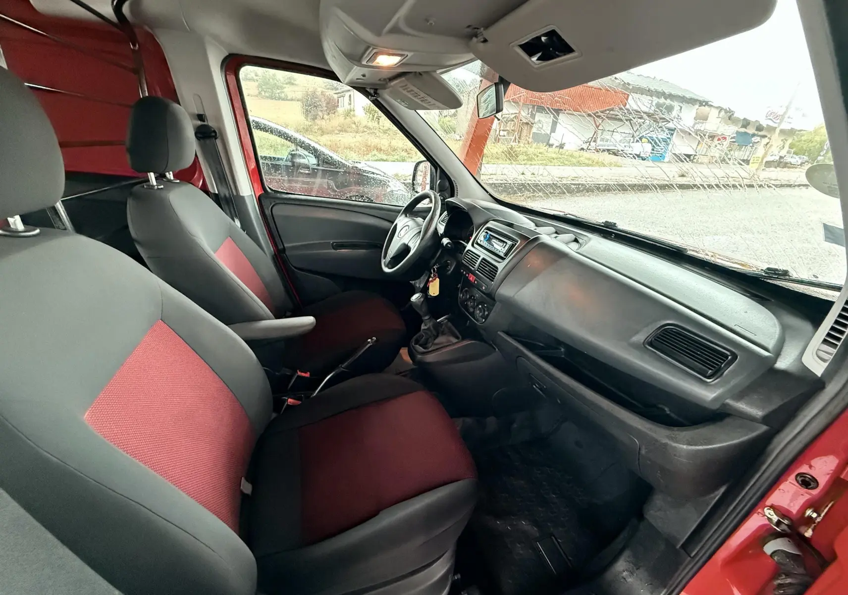 Vue intérieure côté conducteur du Fiat Doblo Cargo rouge de 2011, avec sièges tissu rouge et gris et tableau de bord noir.