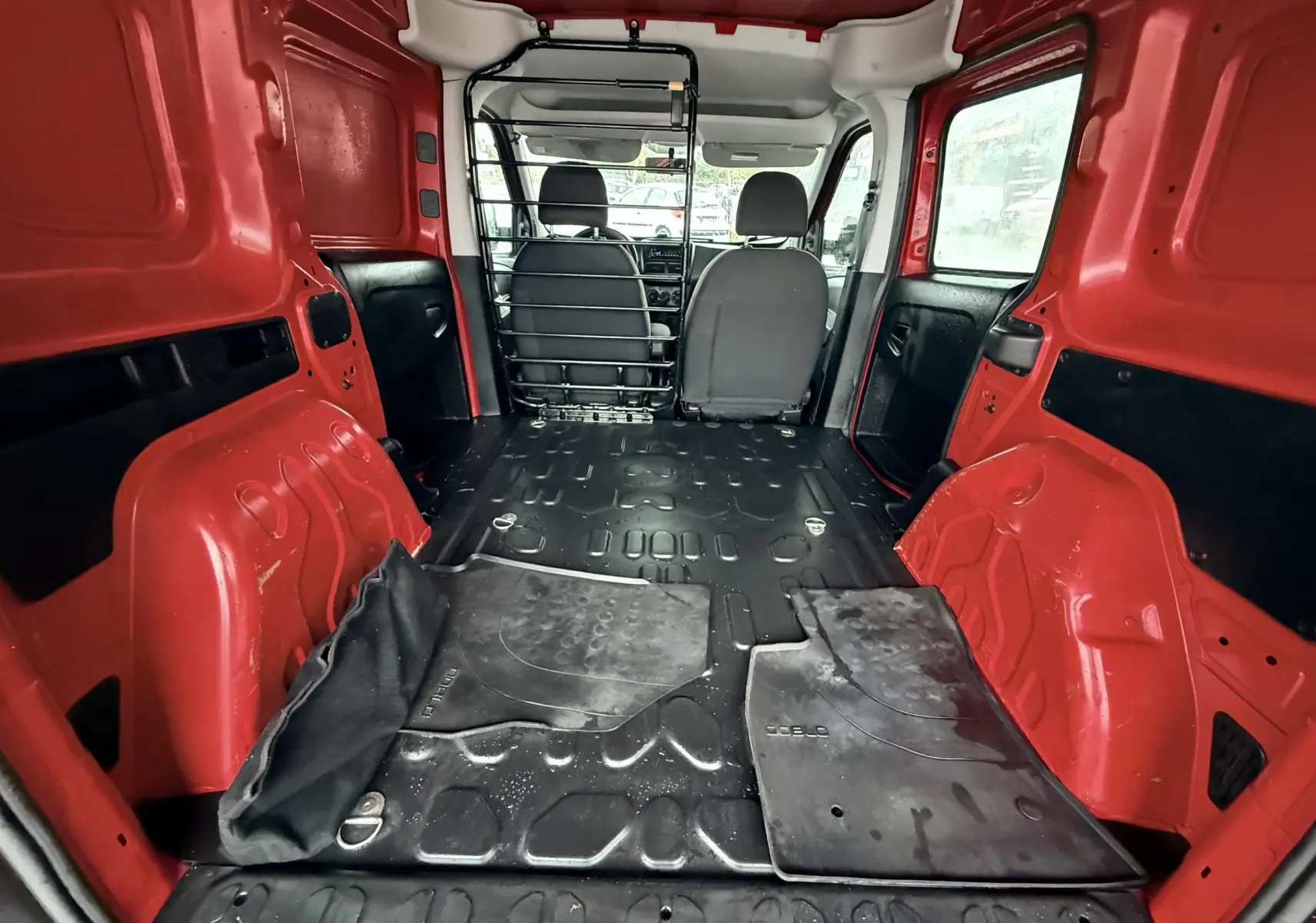 Vue intérieure arrière du FIAT Doblo Cargo rouge, montrant l'espace de chargement vide avec tapis caoutchouc et séparation métallique.
