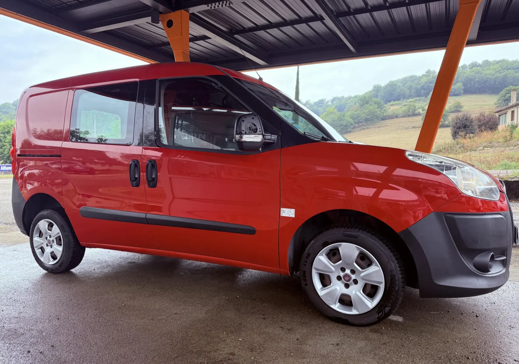 Fiat Doblo Cargo rouge en vue 3/4 avant droit, avec pare-chocs noirs et jantes argentées sous abri.