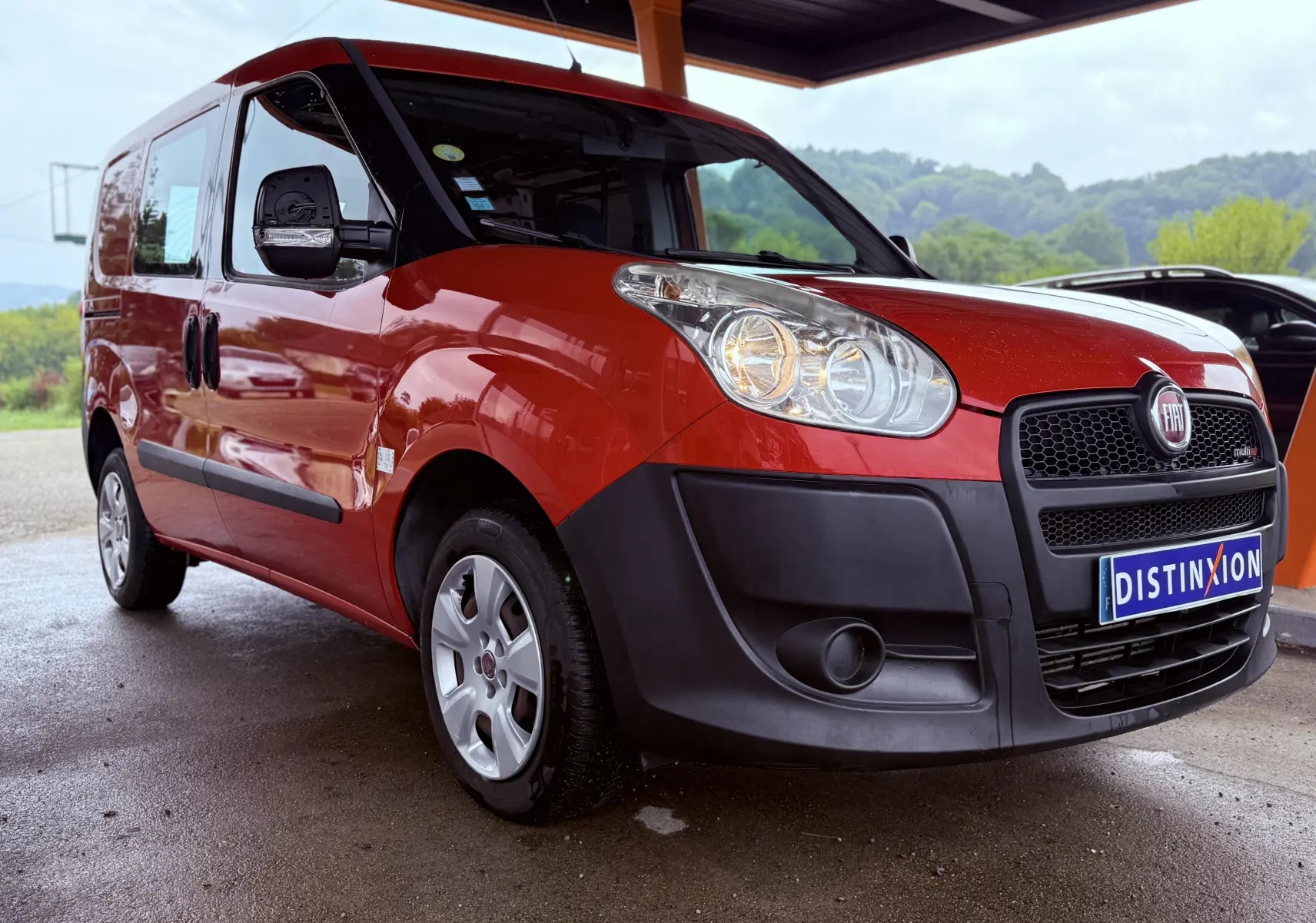 Fiat Doblo Cargo rouge vue 3/4 avant droit sous abri avec pare-chocs noirs et phares allumés.