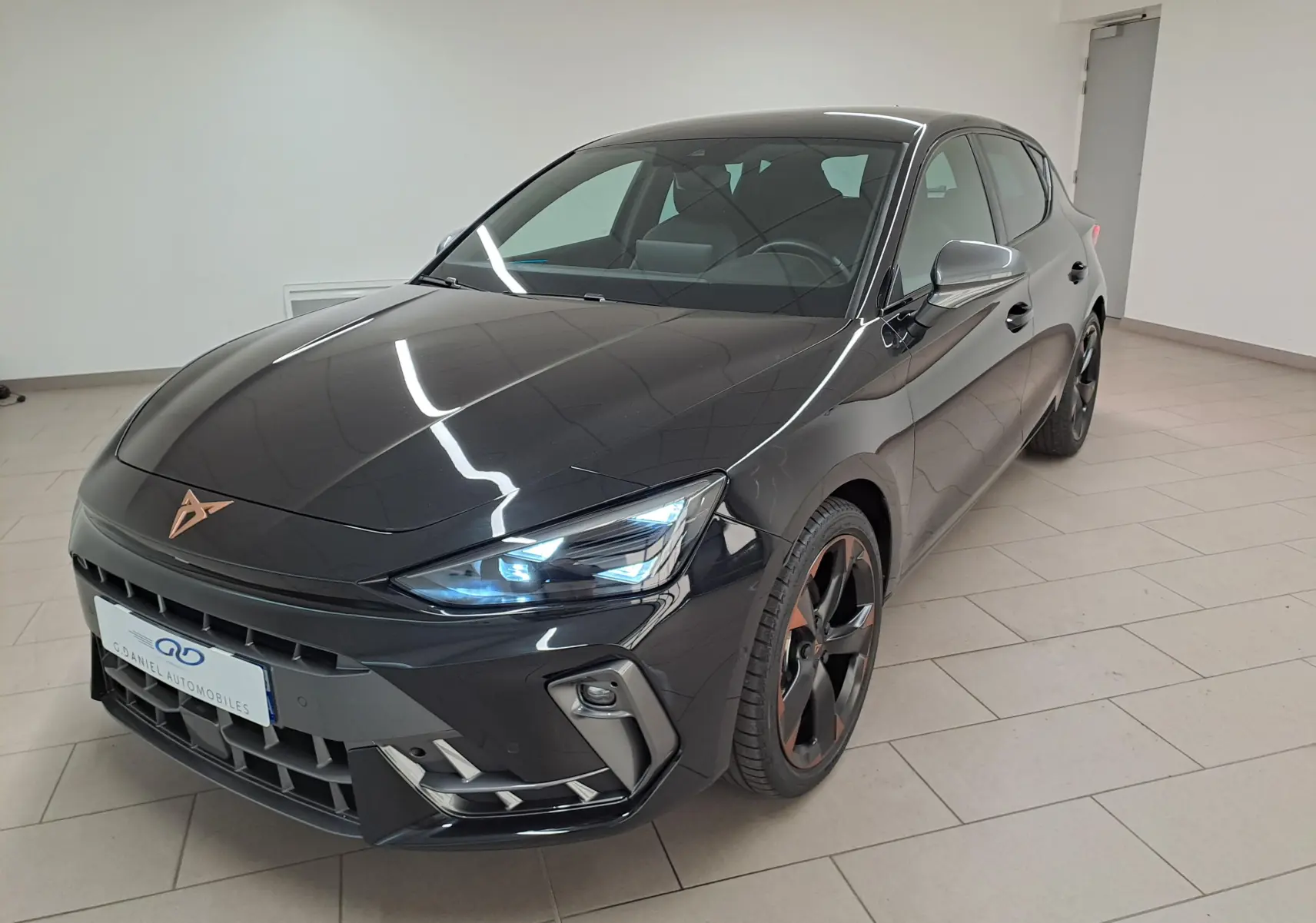 CUPRA Leon Ph.2 noir minuit vu en 3/4 avant droit, avec jantes cuivrées et feux allumés dans un showroom.