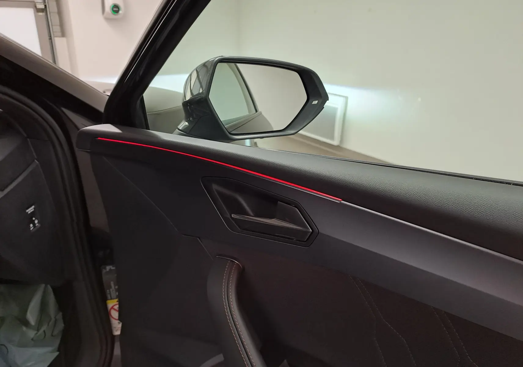 Intérieur côté conducteur de la CUPRA Leon 2025 noir minuit, avec éclairage d'ambiance rouge sur la porte et miroir latéral visible.