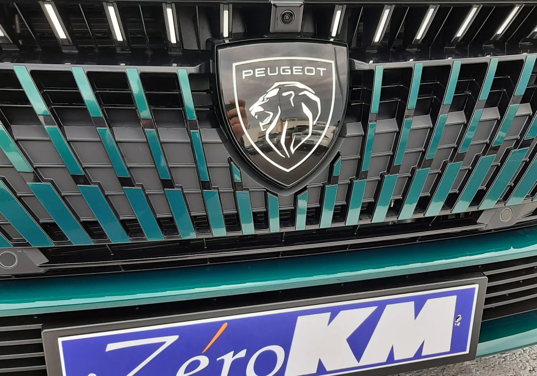 Gros plan sur la calandre bleue Lagoa du Peugeot 308 1.2 Hybrid 145ch GT avec logo lion et plaque Zéro KM.