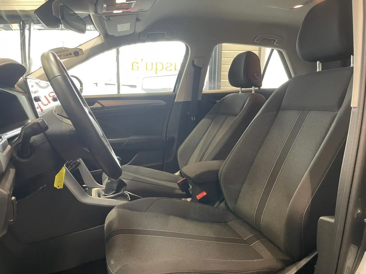 Vue intérieure côté gauche du Volkswagen T-Roc 2025 gris Indium, montrant les sièges avant en tissu noir et le volant multifonction.