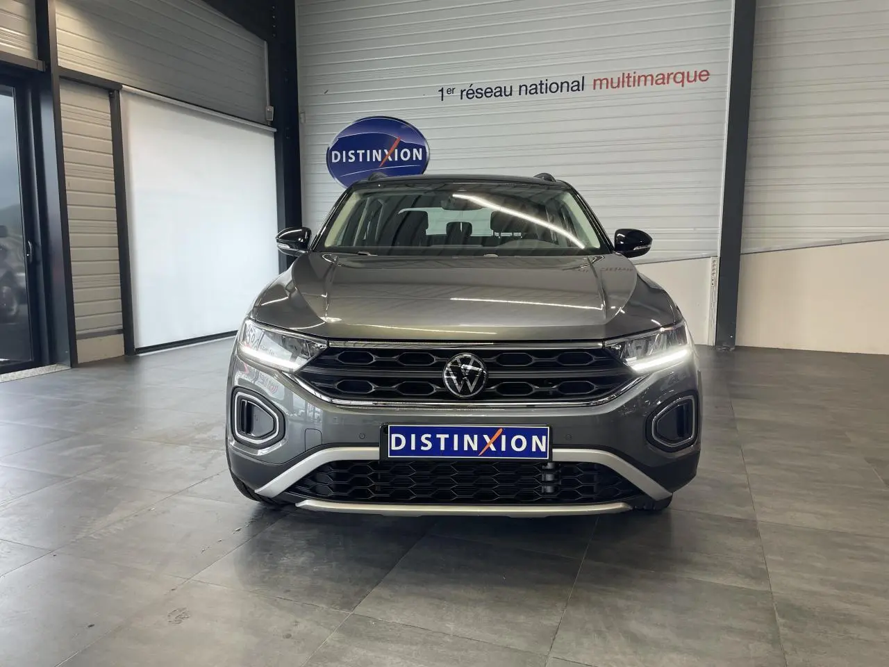 Vue frontale d'un Volkswagen T-Roc 2025 gris Indium avec calandre noire et éclairage LED allumé en intérieur showroom.