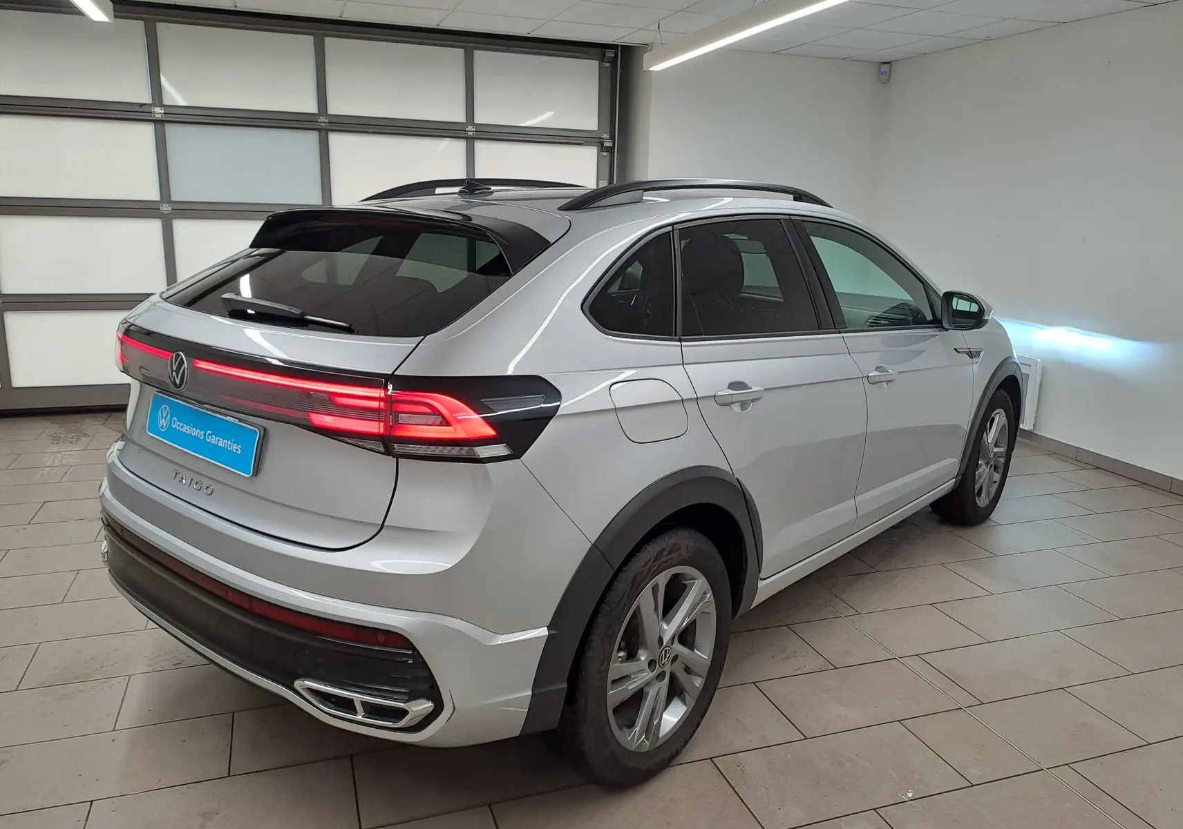 Volkswagen Taigo 2024 R-Line en reflet d'argent, vue 3/4 arrière droit avec feux arrière allumés et toit noir.