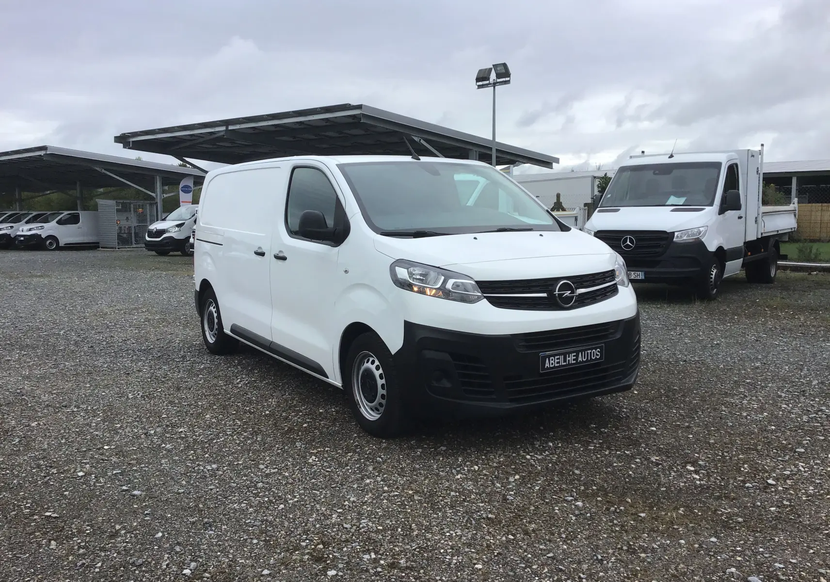 Fourgon Opel Vivaro blanc vu en 3/4 avant droit sur un parking avec d'autres véhicules utilitaires.