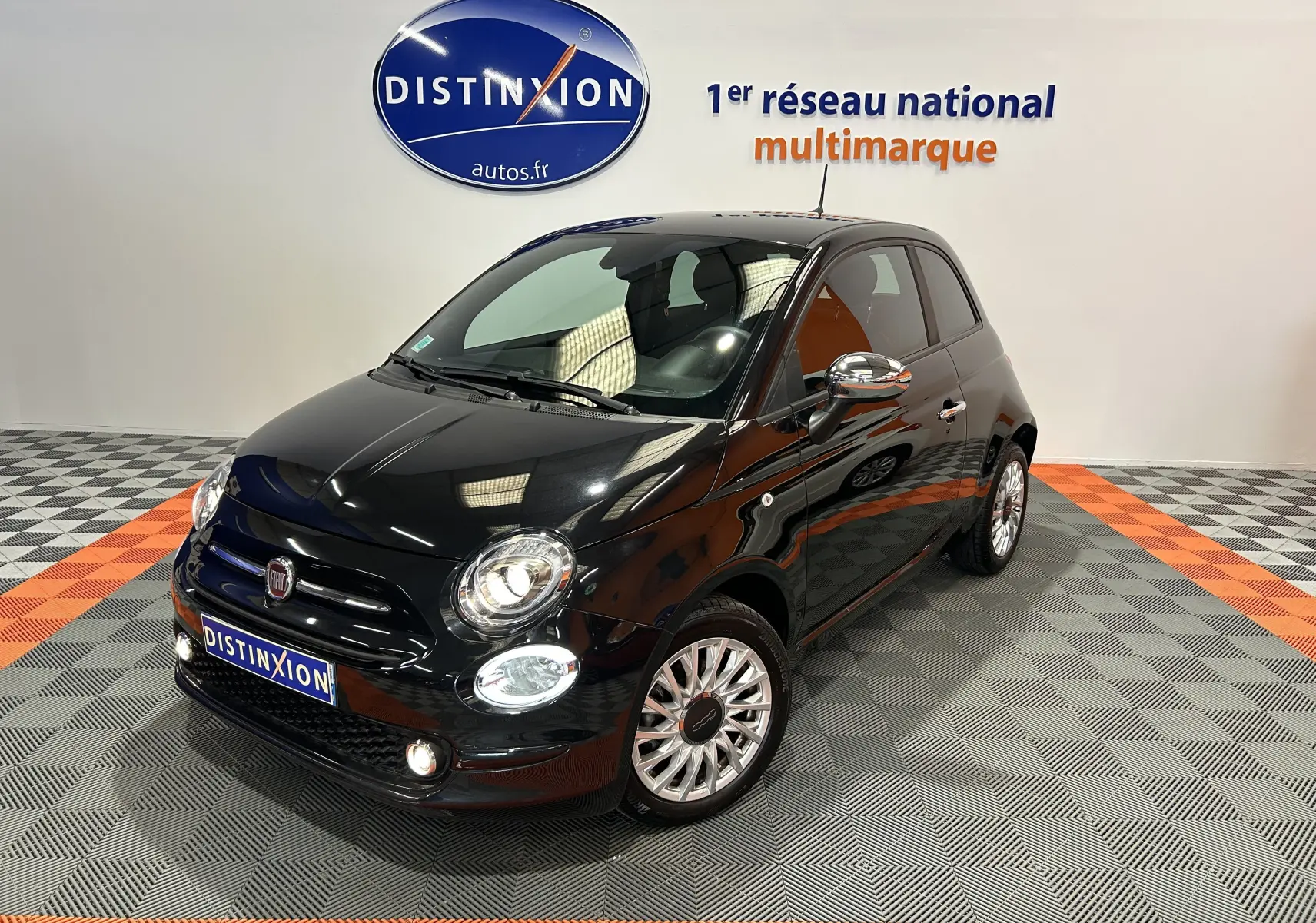 Fiat 500 noire vue 3/4 avant droit dans un showroom avec phares allumés et jantes multibranches.