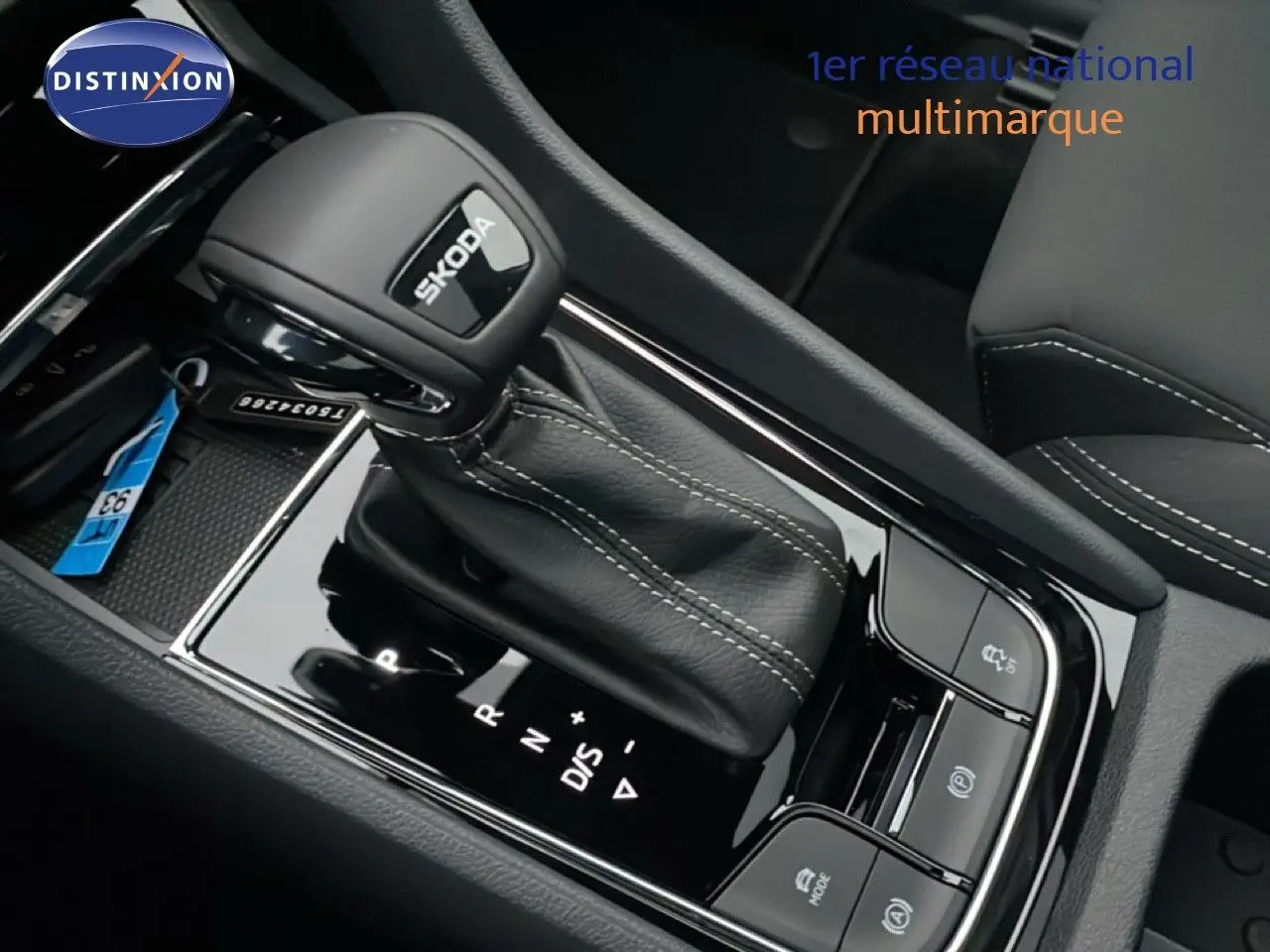Gros plan sur le levier de vitesse automatique noir avec logo SKODA, entouré de commandes et coutures blanches visibles.