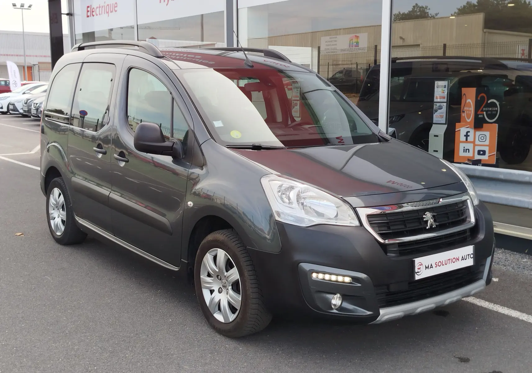 Peugeot Partner Tepee gris foncé vu en 3/4 avant droit, avec barres de toit et feux diurnes allumés.