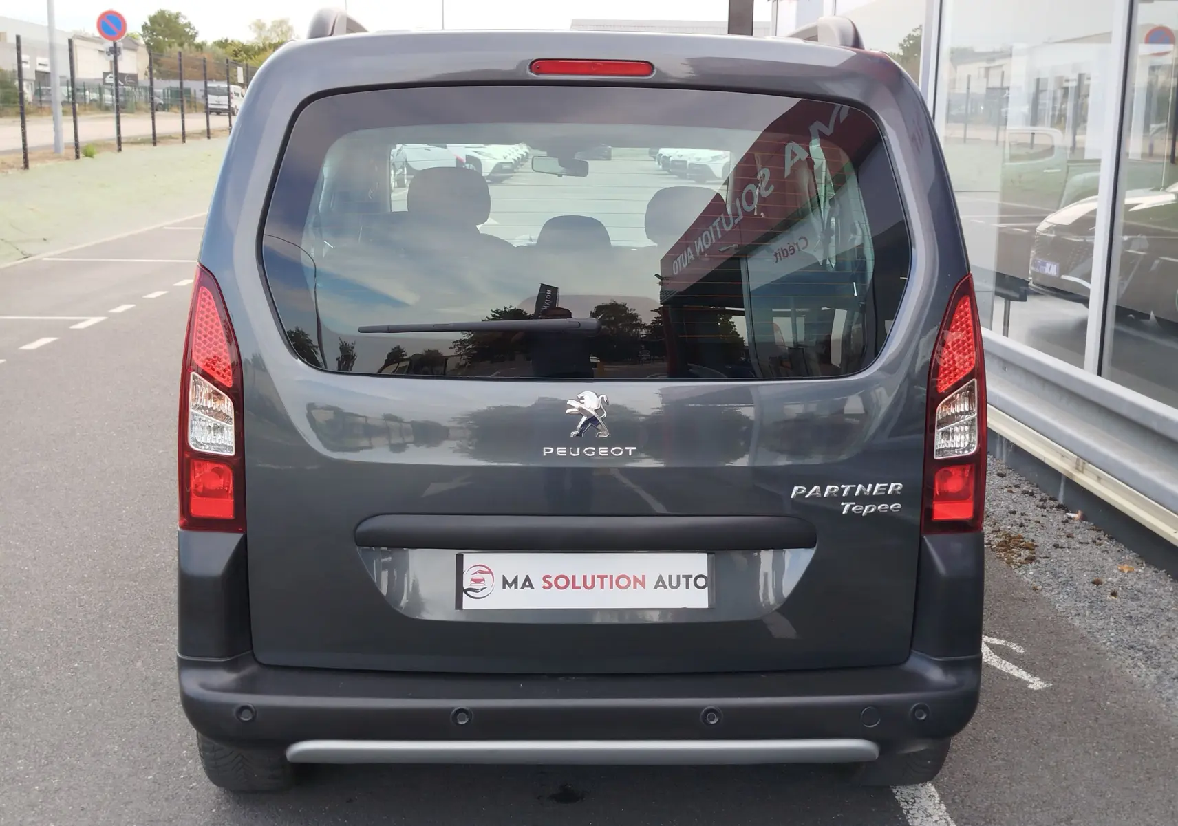 Vue arrière d'un Peugeot Partner Tepee gris foncé, avec logo et badges visibles, stationné devant un bâtiment vitré.