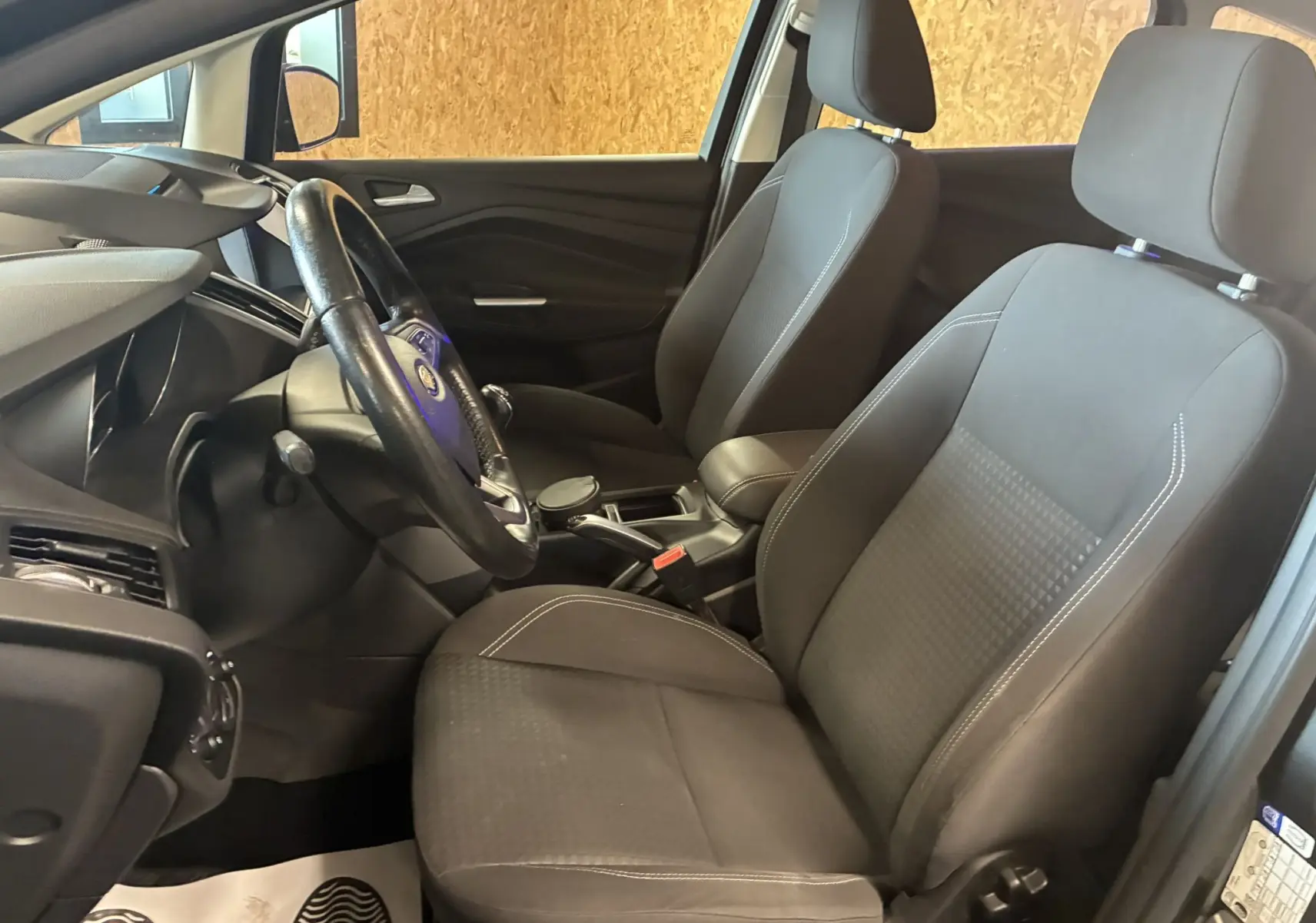 Vue intérieure côté conducteur du Ford C-MAX 2015, sièges tissu gris noir et volant cuir avec levier de vitesses manuel.