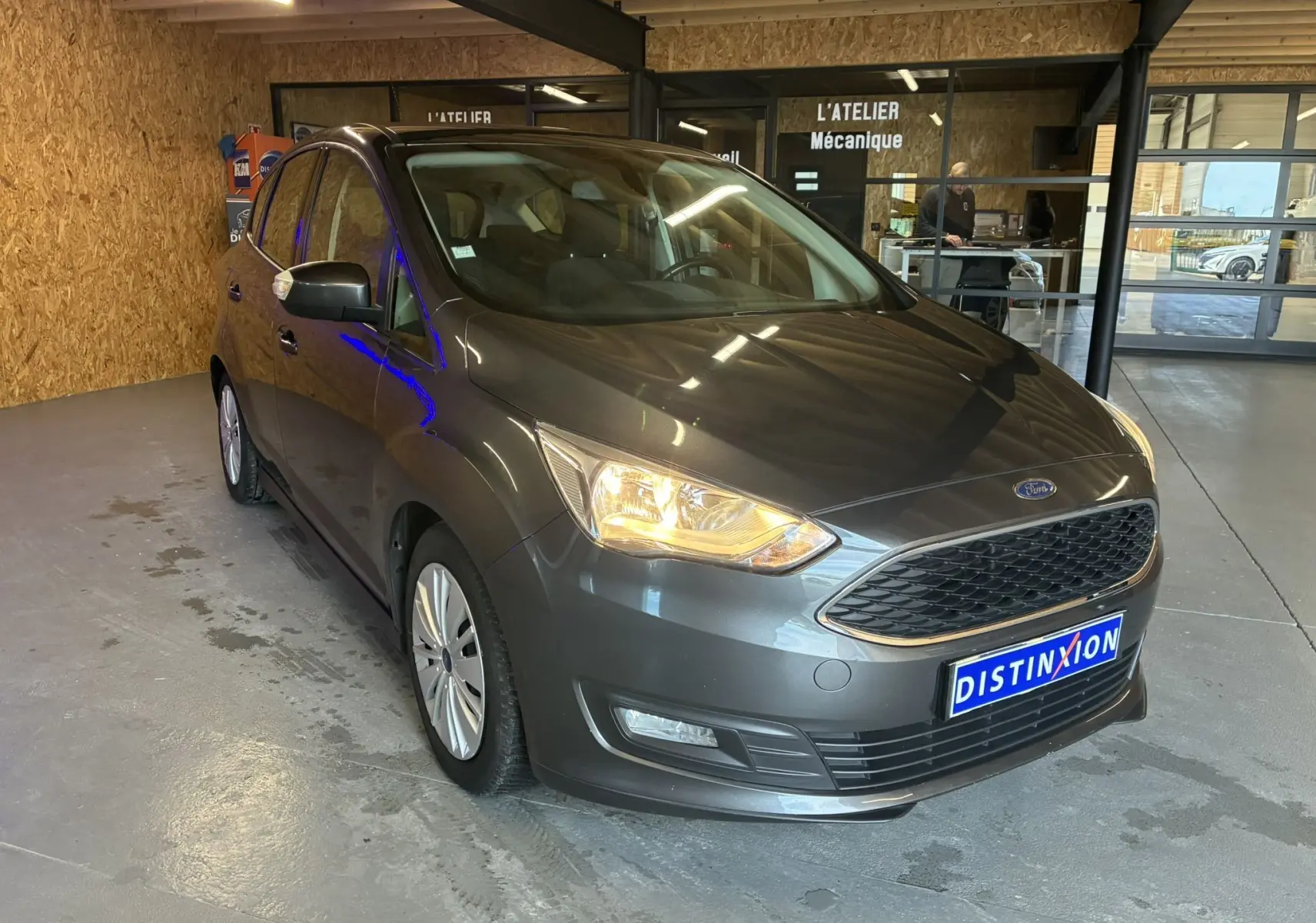 Ford C-MAX gris en 3/4 avant droit dans un garage avec phare avant allumé et plaque Distinxion visible.