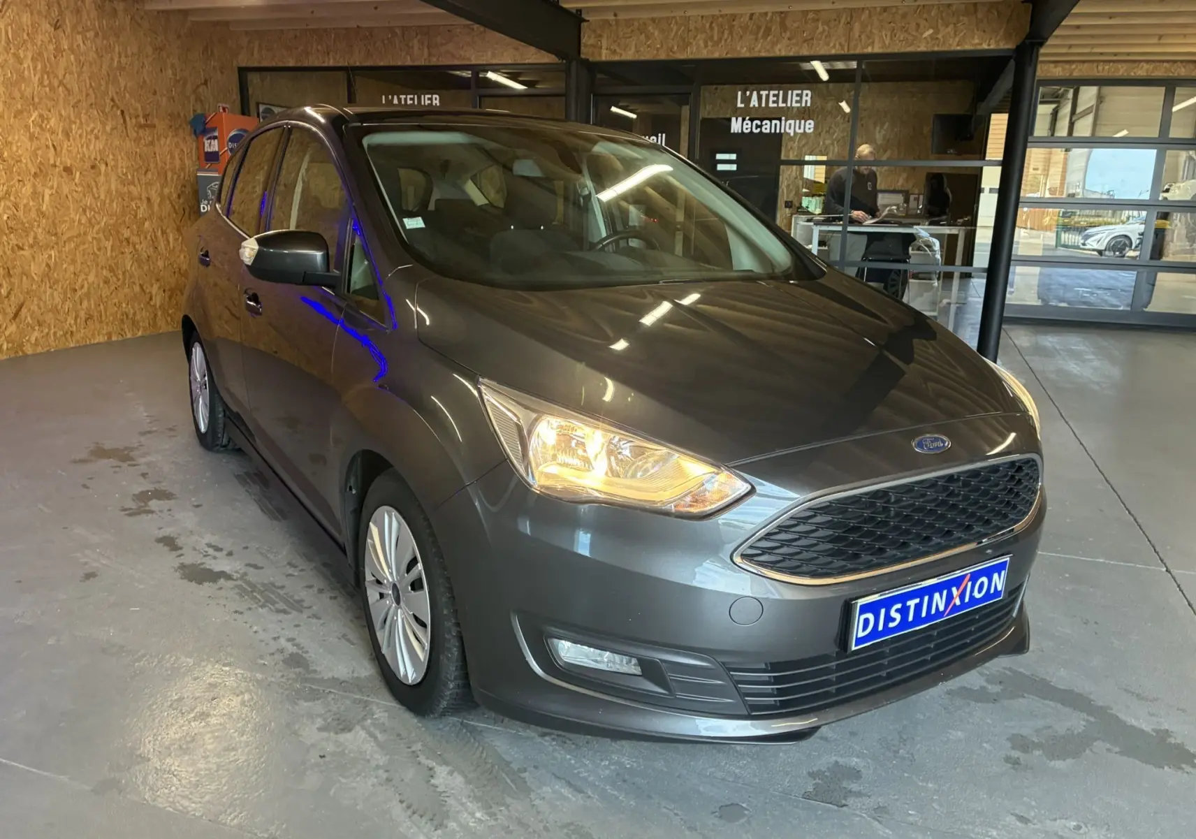 Ford C-MAX gris en 3/4 avant droit avec phares allumés, garée dans un atelier mécanique intérieur.