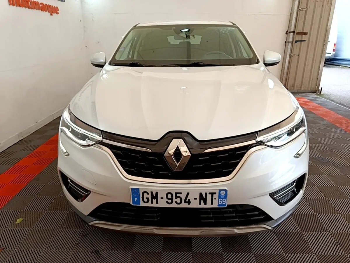 Vue frontale d'une Renault Arkana blanche 2023 avec calandre noire et phares LED allumés dans un garage.