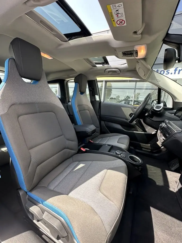 Intérieur de la BMW I3 2022 vue côté conducteur, sièges gris avec surpiqûres bleues et toit ouvrant panoramique.