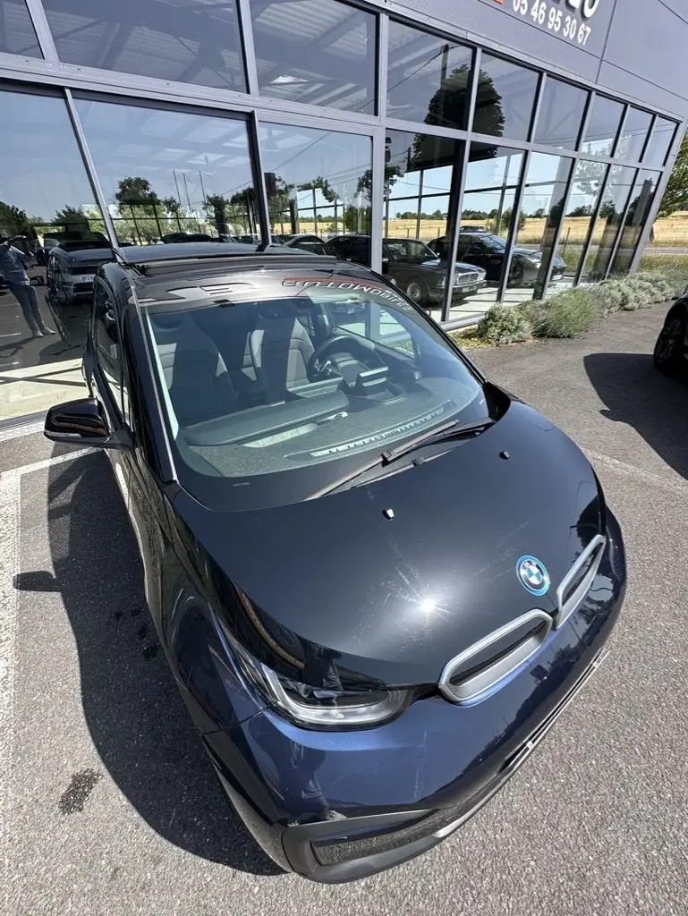 BMW i3 électrique bleue vue en 3/4 avant avec toit noir brillant et logo BMW visible sur le capot.