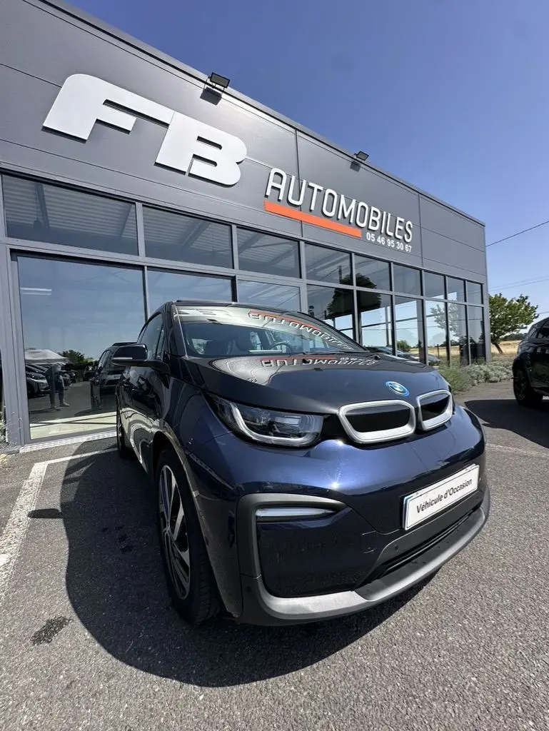 BMW i3 bleue vue en 3/4 avant droit, garée devant un concessionnaire FB Automobiles par temps clair.