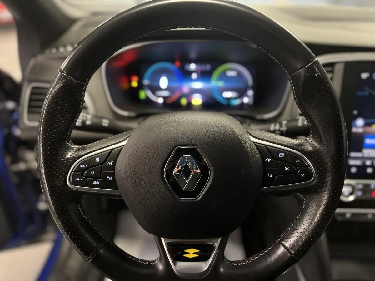 Volant cuir perforé Renault Mégane 1.6 E-Tech bleu, avec commandes intégrées et tableau de bord numérique flou en arrière-plan.