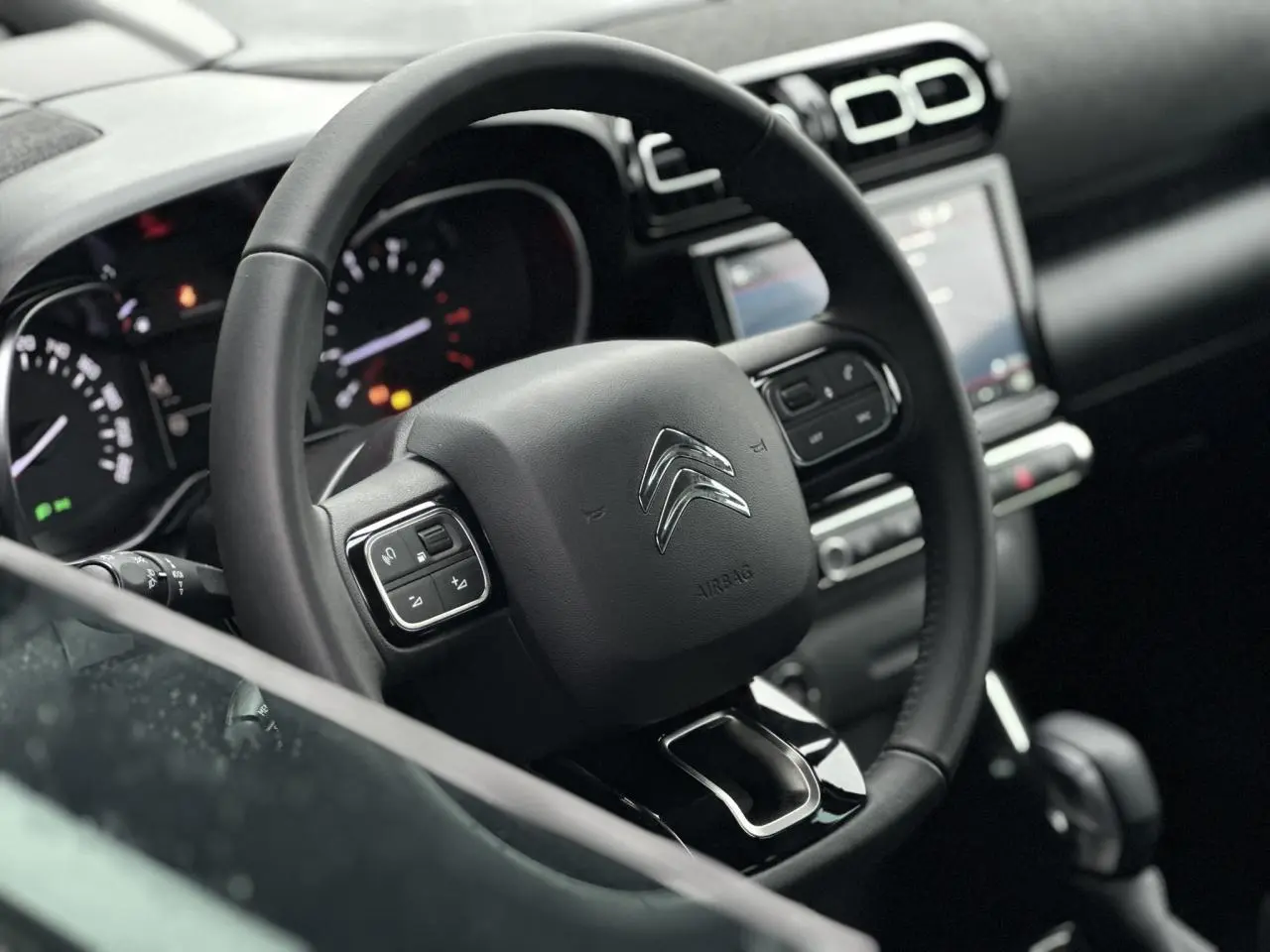 Vue rapprochée du volant noir de la Citroën C3 Aircross 2024 avec commandes intégrées et tableau de bord en arrière-plan.