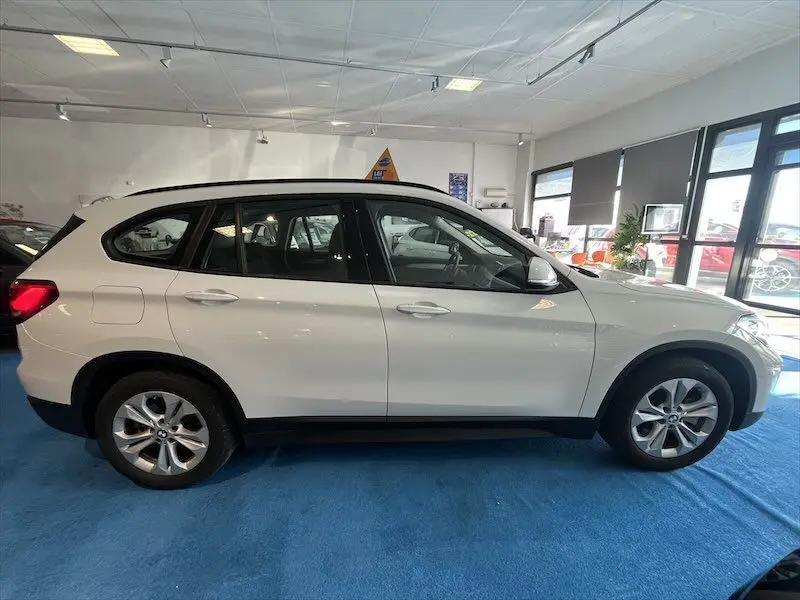 Profil côté gauche d'un BMW X1 XDRIVE blanc alpin exposé en intérieur sur sol bleu.