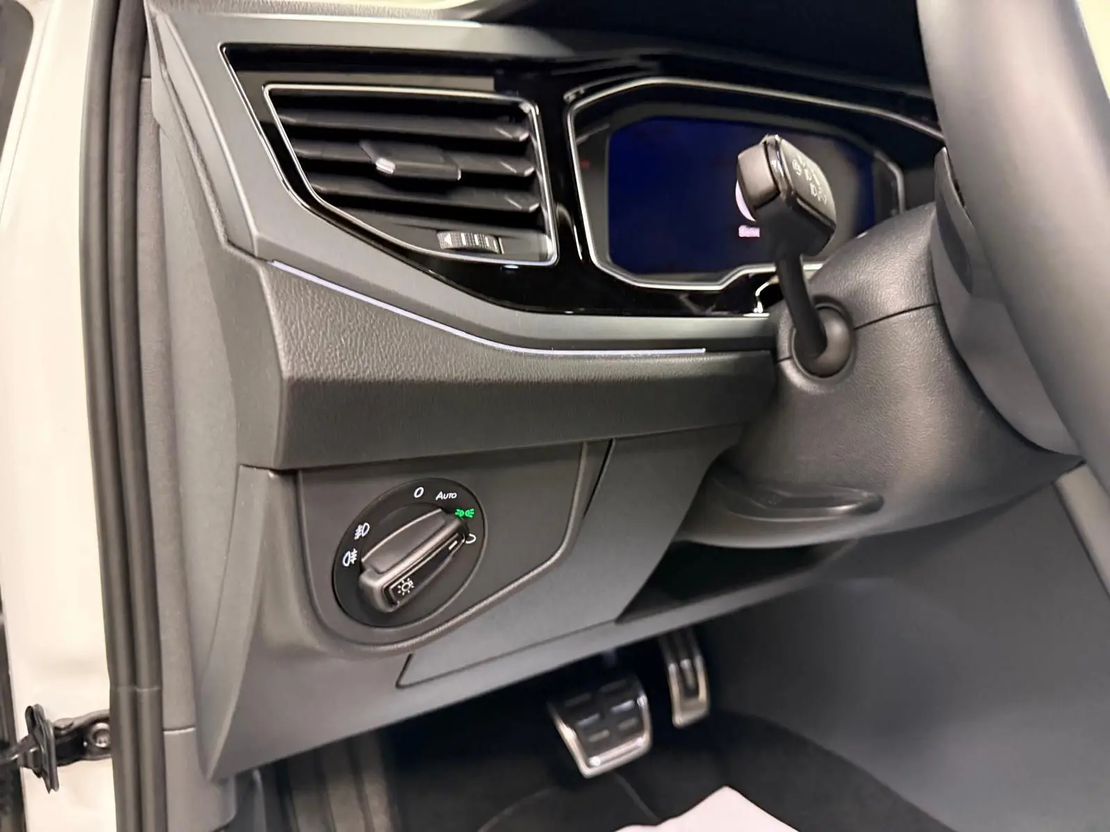 Détail intérieur côté gauche du tableau de bord du Volkswagen Taigo blanc, avec commande des phares et pédalier en acier.