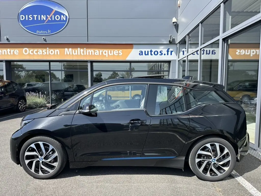 Profil côté gauche d'une BMW i3 noire électrique 2022, avec jantes distinctives et toit vitré, stationnée devant un showroom.