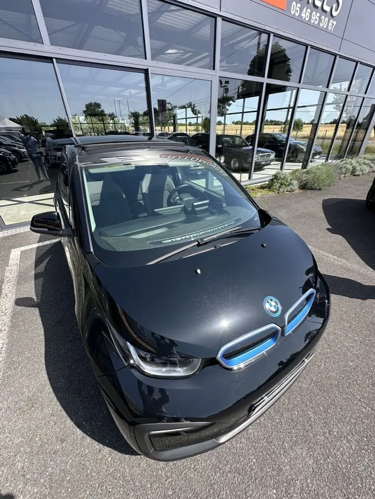 Vue 3/4 avant d'une BMW i3 noire électrique 2022, avec calandre bleue distinctive et pare-brise reflétant un bâtiment vitré.
