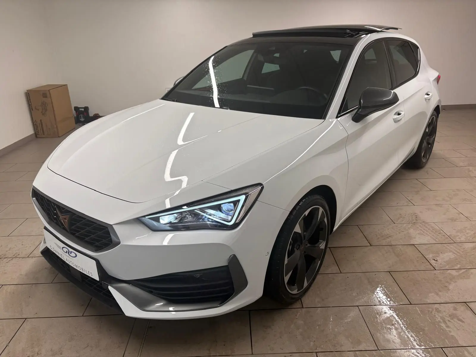 Vue 3/4 avant droite de la CUPRA Leon blanche Nevada avec toit ouvrant panoramique et jantes noires en intérieur.