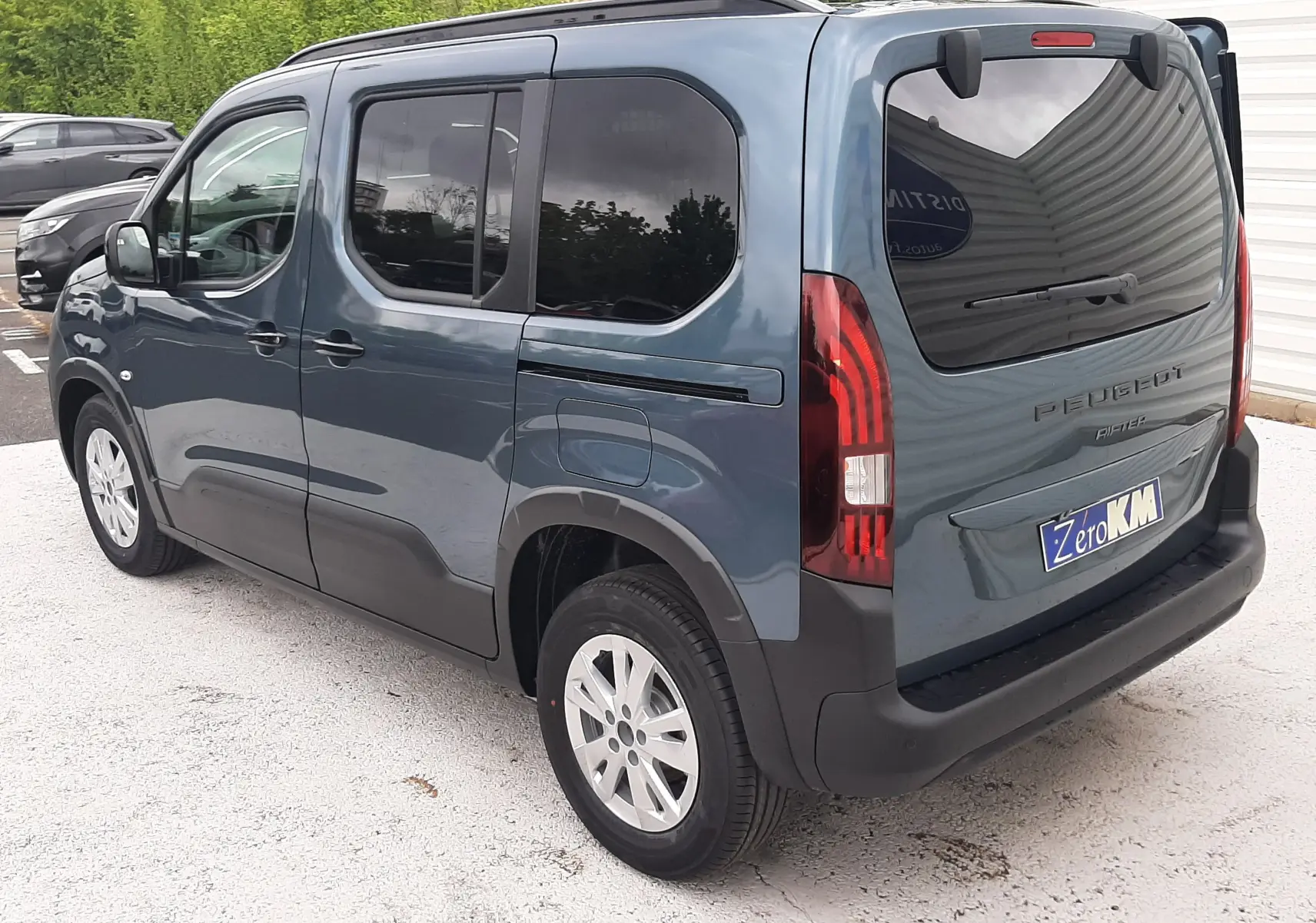 Vue 3/4 arrière droite d'un Peugeot Rifter bleu Kiama 2026 avec vitres teintées et hayon fermé sur parking.