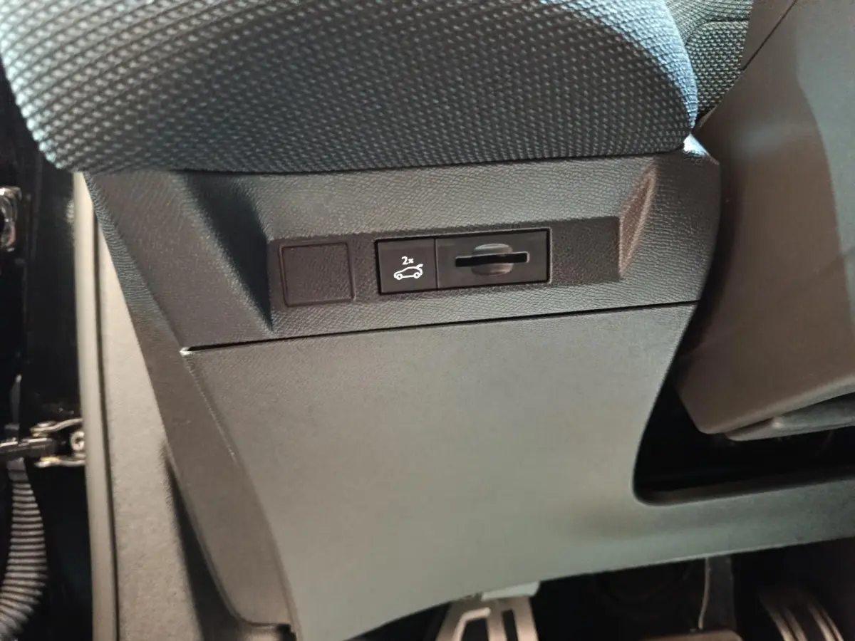 Gros plan sur le bouton d'ouverture du coffre noir situé sous le siège avant du Citroën C5 Hybride 2026.