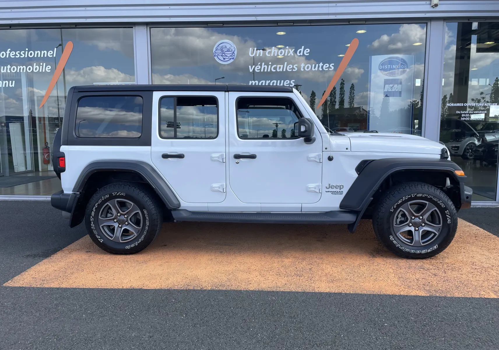 Jeep Wrangler blanc vu de profil côté gauche, avec toit noir et jantes gris foncé sur fond de vitrine commerciale.