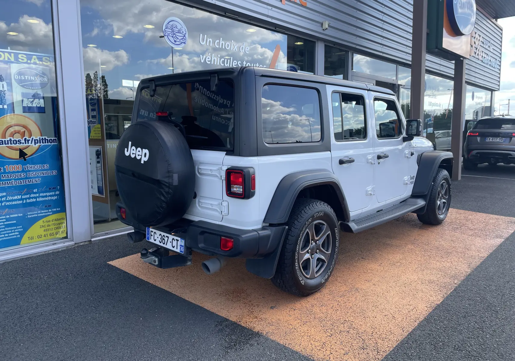 Vue 3/4 arrière droite d'un Jeep Wrangler blanc avec roue de secours noire et toit rigide foncé stationné devant un garage.