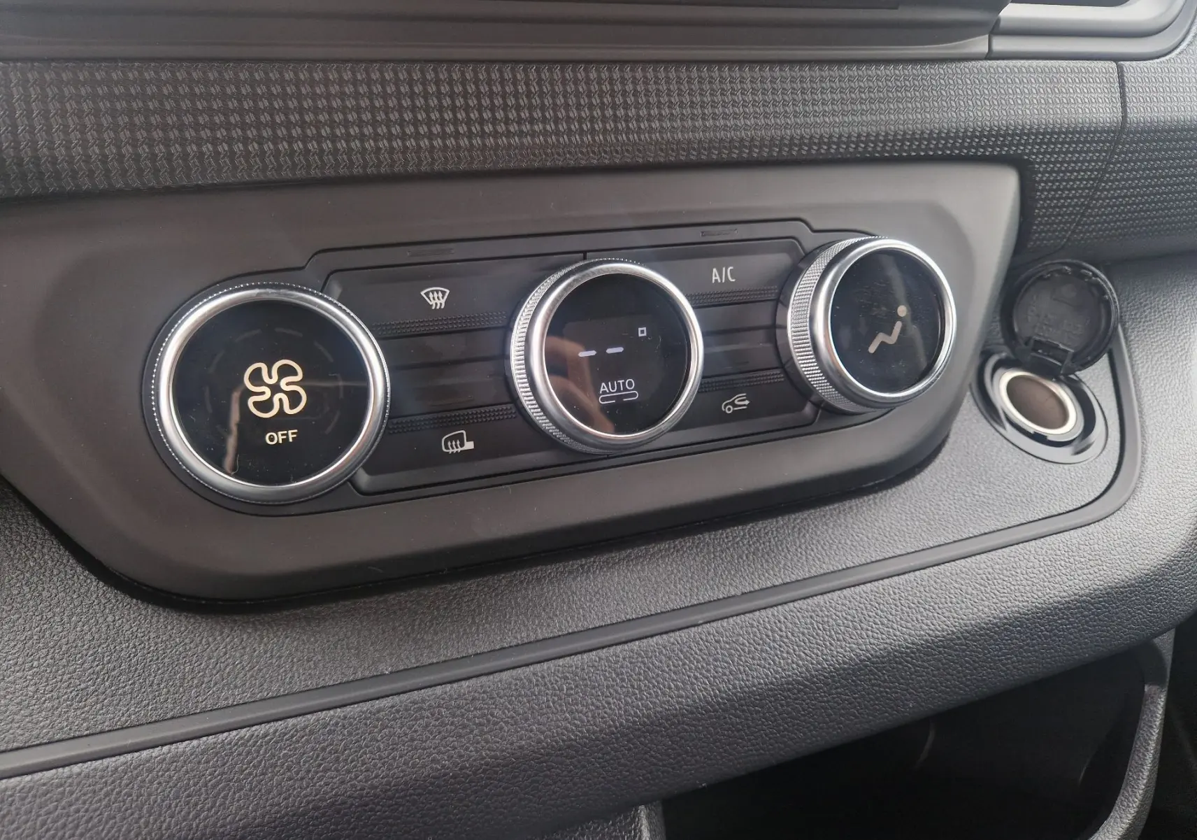 Gros plan sur la console de climatisation du Renault Trafic Fourgon gris urbain, montrant les trois boutons rotatifs et une prise 12V ouverte.