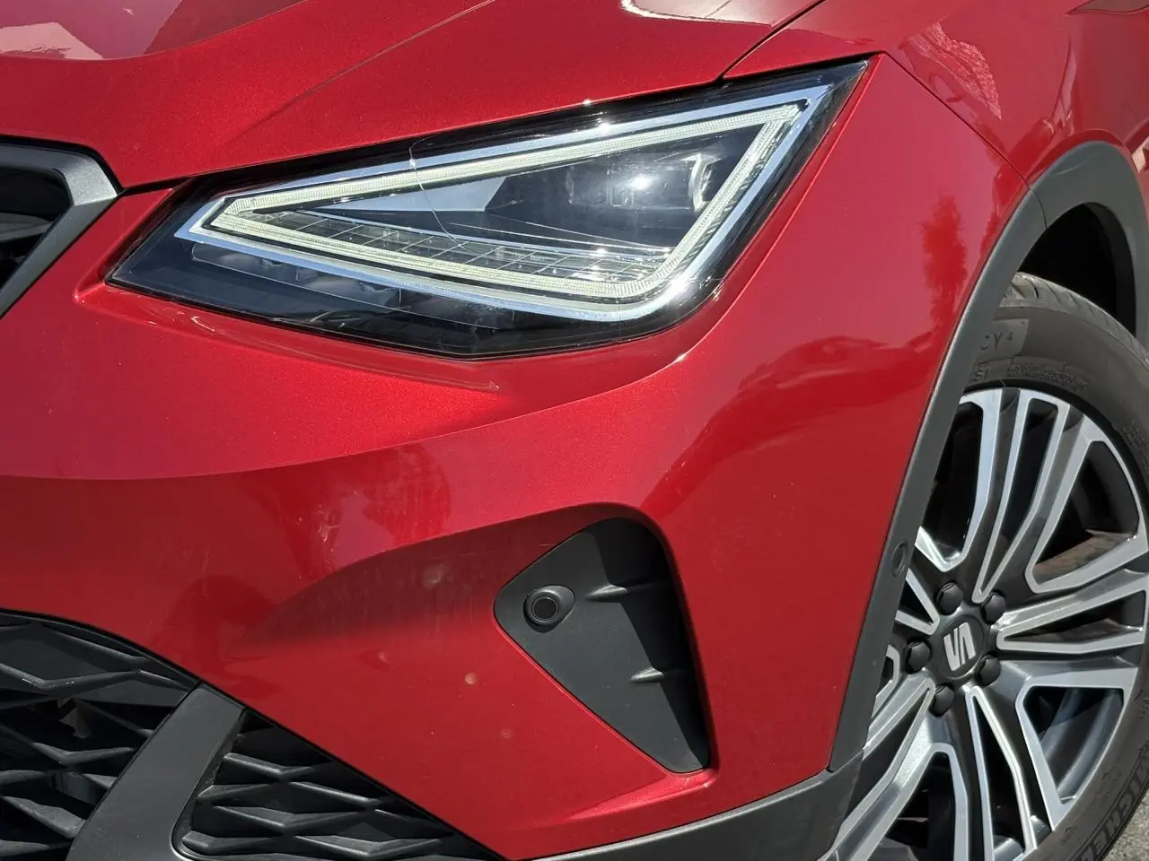 Gros plan sur le phare avant gauche rouge Désir du SEAT ARONA TSI 110 FR avec jante alliage 17 pouces et caméra frontale.