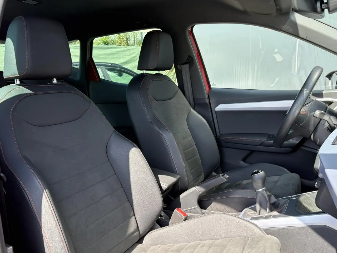 Intérieur avant du SEAT Arona TSI 110 FR 2024, sièges en suédine noire avec surpiqûres rouges, volant et levier de vitesse visibles.