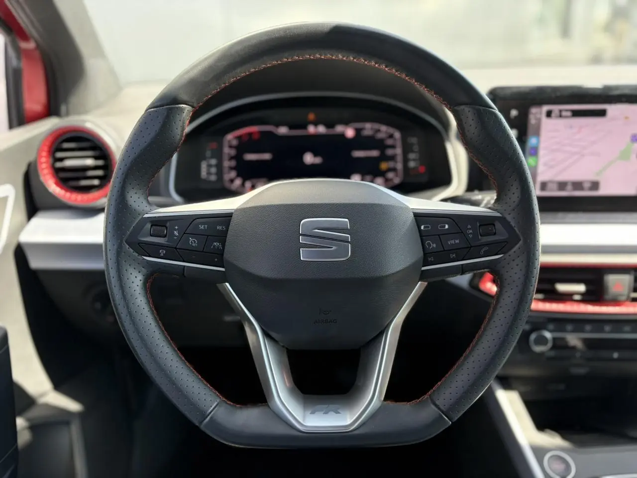 Volant sport noir avec surpiqûres rouges du SEAT Arona TSI 110 FR 2024, tableau de bord digital et écran tactile en arrière-plan.