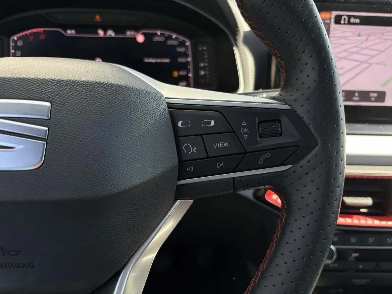 Gros plan sur le volant perforé noir avec surpiqûres rouges du SEAT Arona TSI 110 FR, tableau de bord numérique en arrière-plan.
