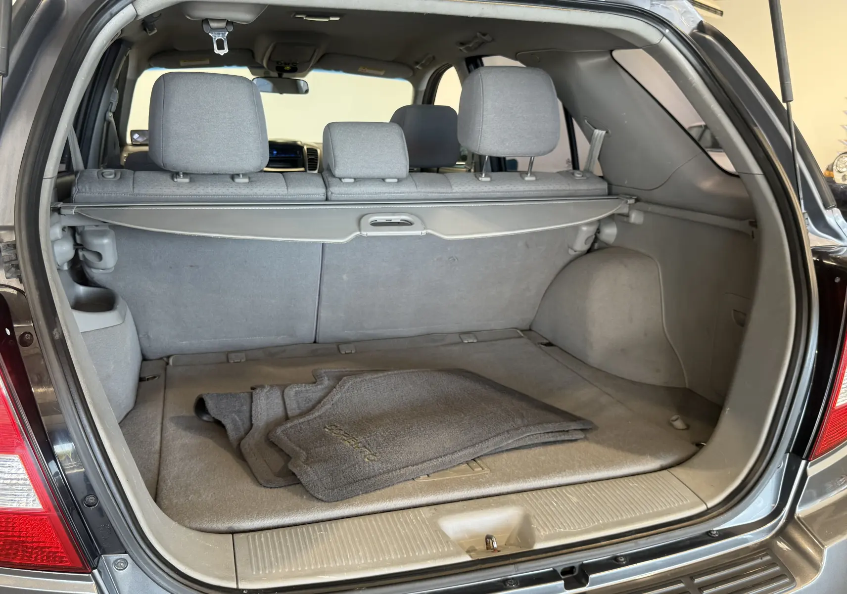 Coffre ouvert d'un KIA Sorento gris 2005, vue arrière montrant tapis de sol gris et banquette arrière rabattue partiellement.