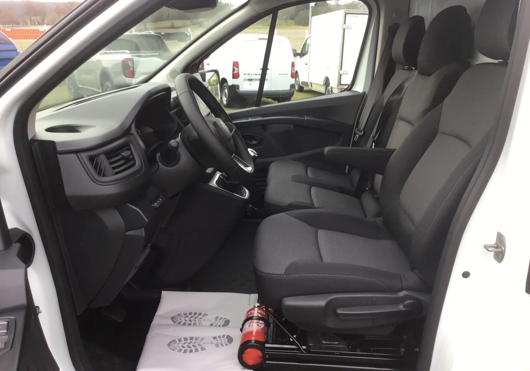 Vue intérieure côté conducteur du Renault Trafic Fourgon blanc 2025, montrant les sièges tissu gris et le volant.