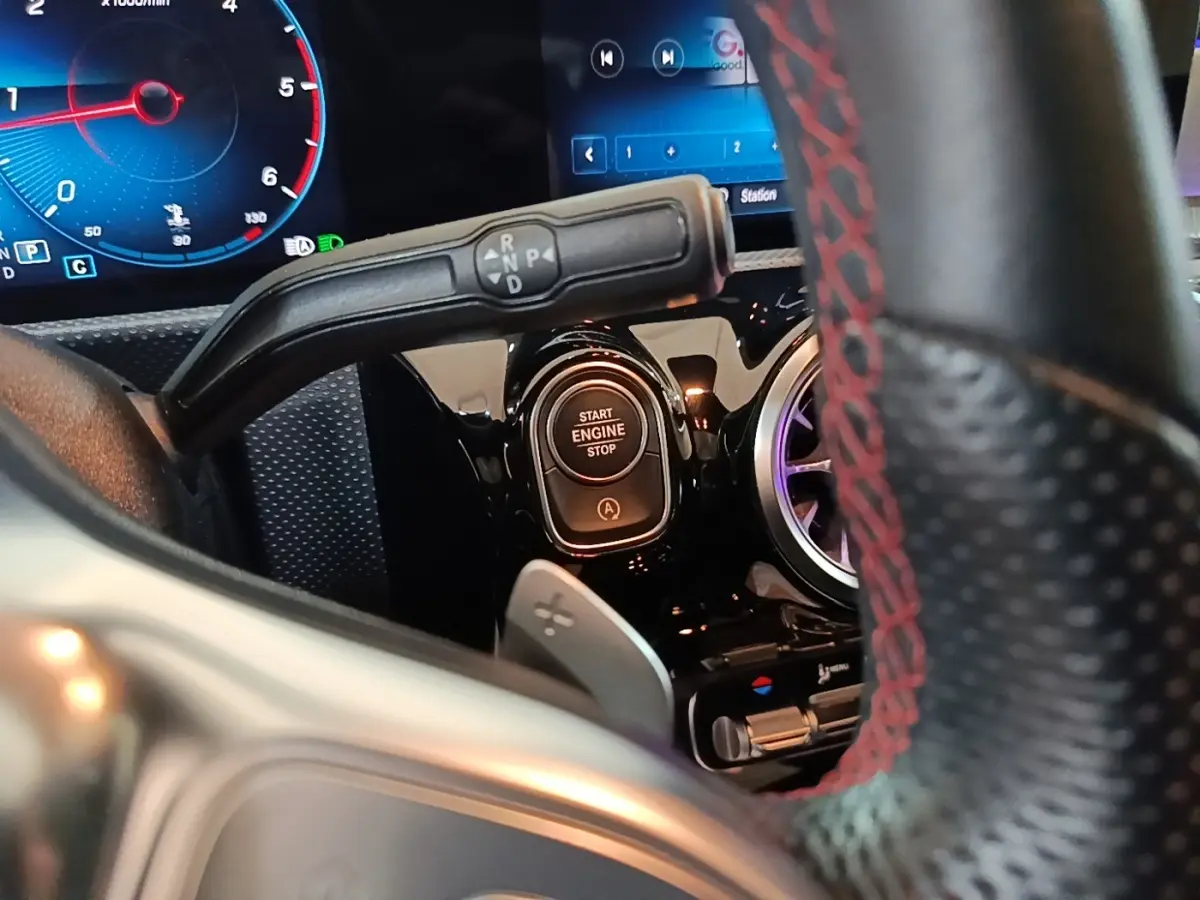 Gros plan intérieur sur le bouton démarrage et levier de boîte automatique d’une Mercedes CLA 200 d AMG Line noire, volant cuir noir à surpiqûres rouges visible.